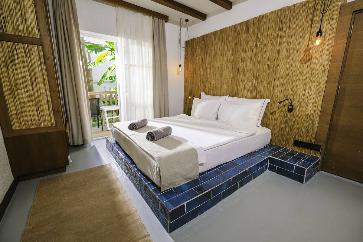 Une chambre à coucher avec un lit, une tête de lit en bois, une fenêtre avec des rideaux et une vue sur la verdure.