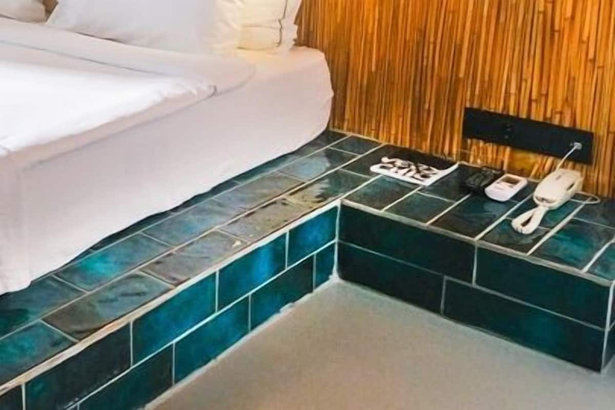 Un lit bien fait, avec deux serviettes enroulées, une table de chevet avec un téléphone et un chargeur, et un sol carrelé.