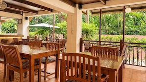 A wooden dining area with tables and chairs, large windows, and a view of greenery outside.