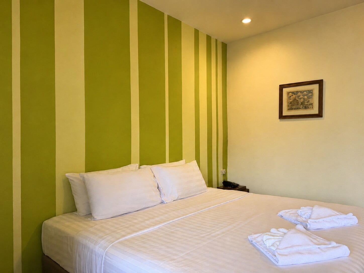Habitación de hotel con una cama, almohadas, una mesita de noche, teléfono y un cuadro en la pared.