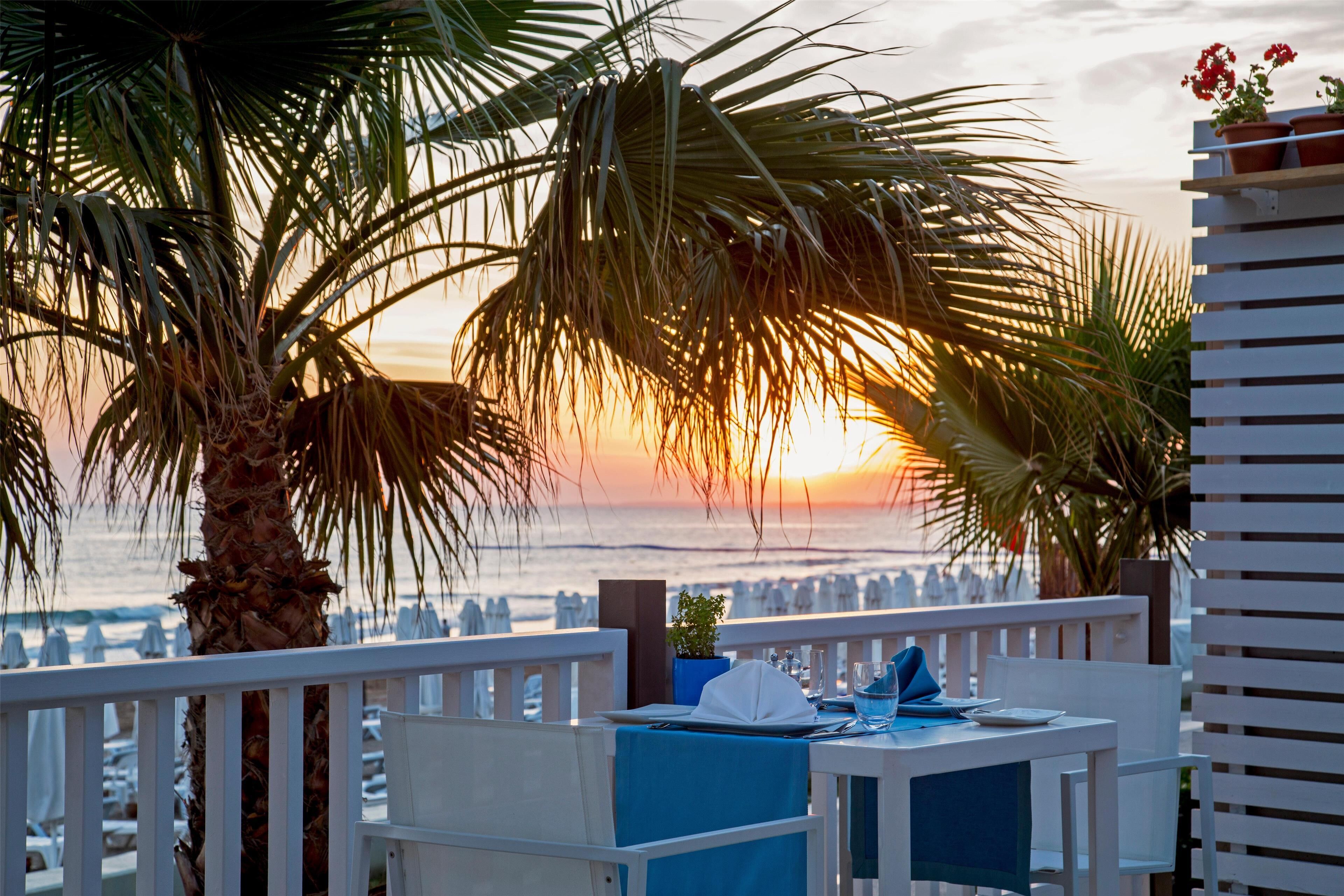 A terrace with a table set for dining, overlooking a sunset over the sea.