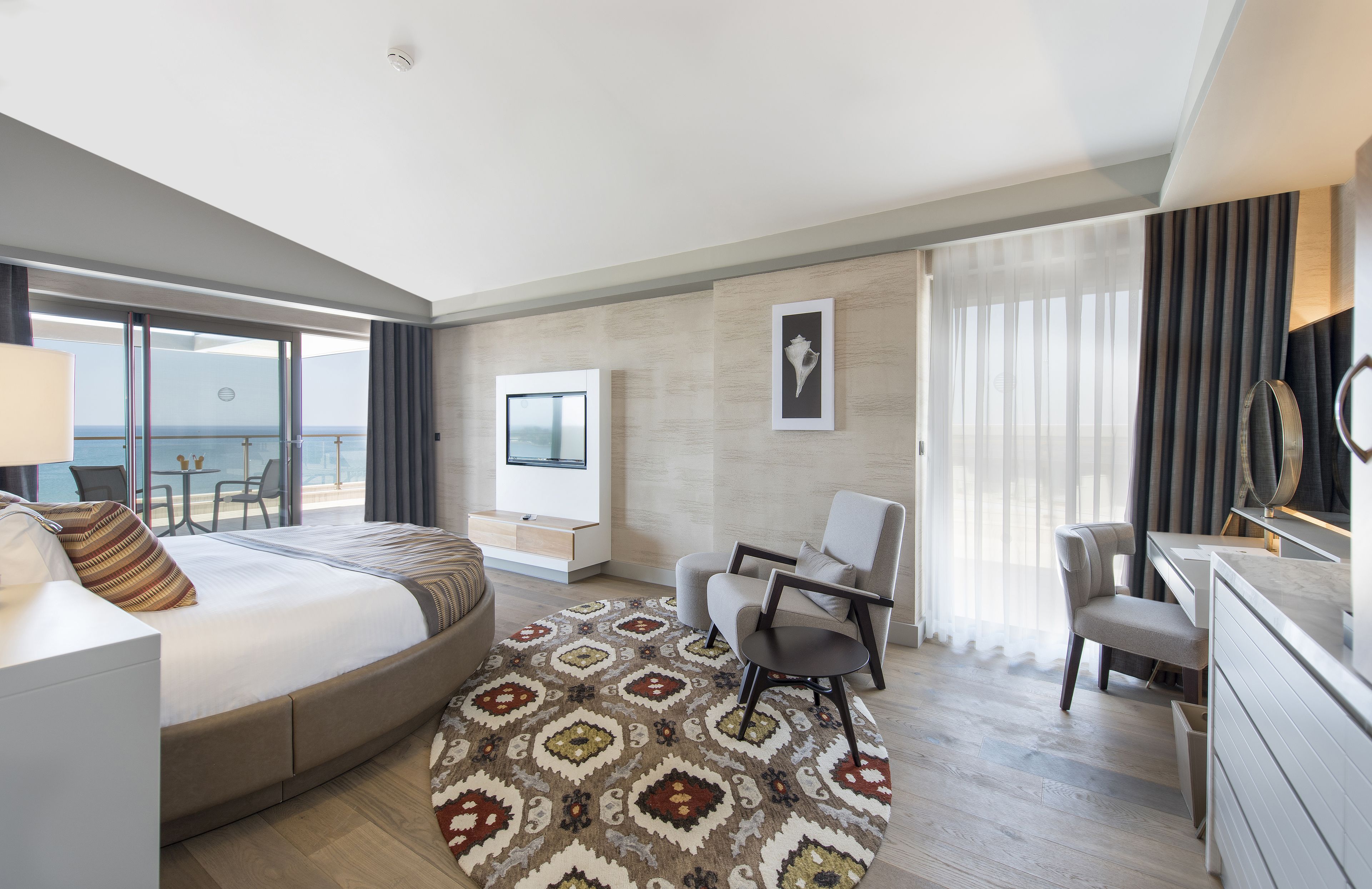 A modern hotel room with a large bed, a desk, a chair, a TV, and a balcony with a view of the sea.