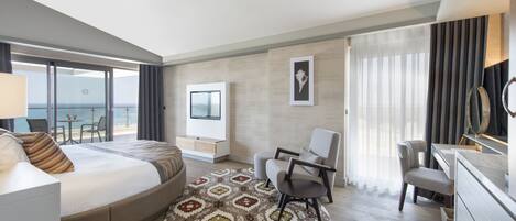 A modern hotel room with a large bed, a desk, a chair, a TV, and a balcony with a view of the sea.
