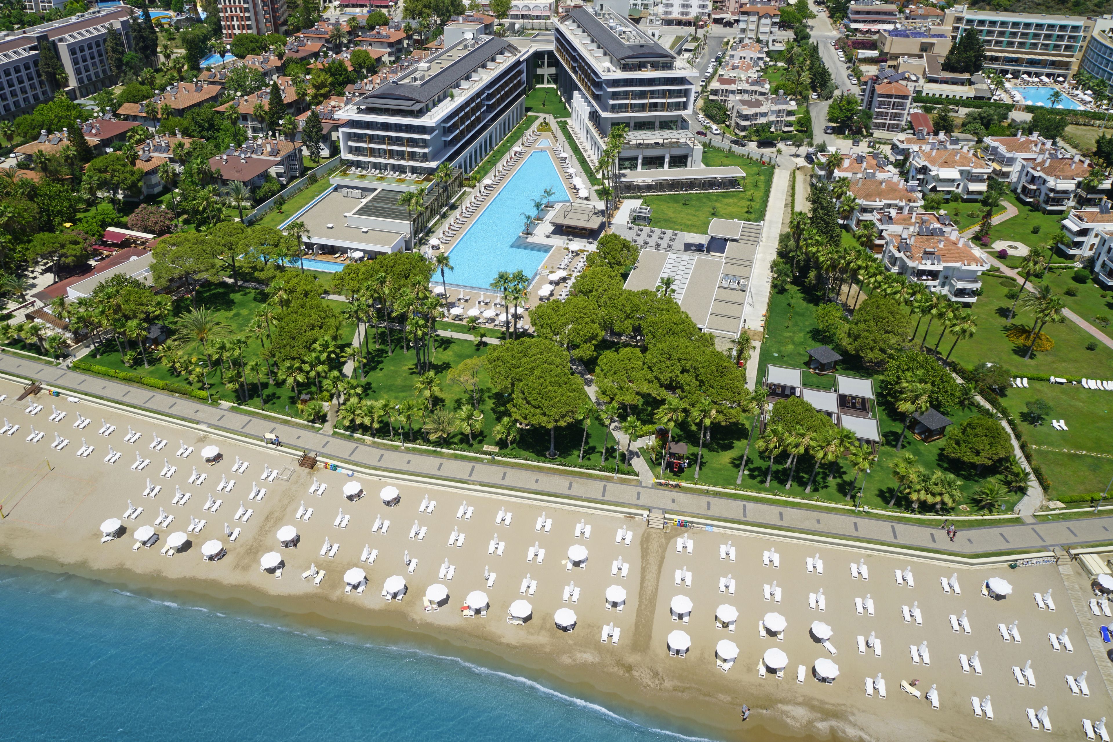 Eine Vogelperspektive zeigt einen Urlaubsort mit großem Pool, Strand mit Liegestühlen und modernen Gebäuden.