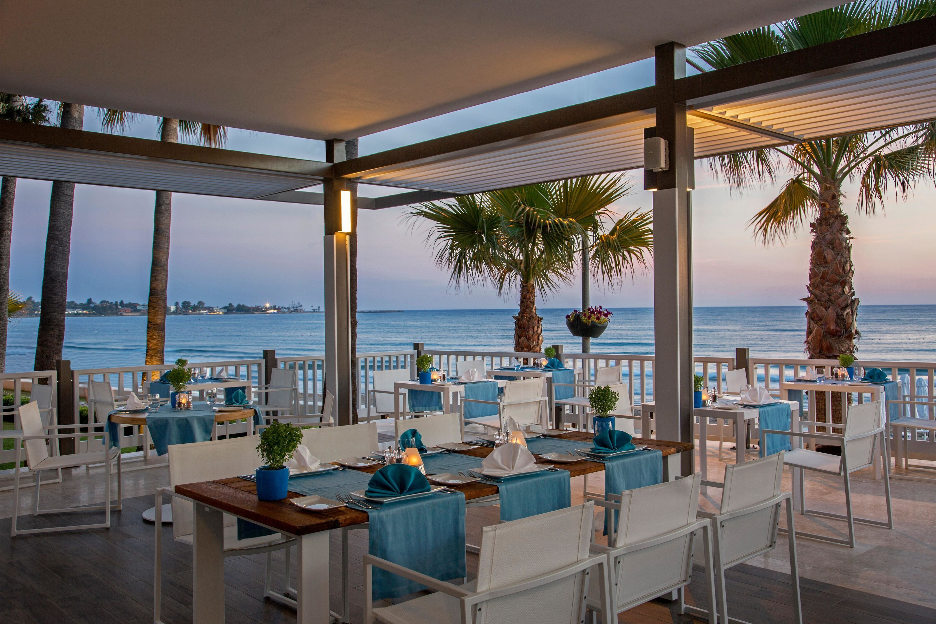 Un restaurante con terraza al aire libre con vistas al mar, con mesas de manteles azules y sillas blancas.