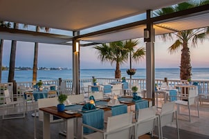Un restaurante con terraza al aire libre con vistas al mar, con mesas de manteles azules y sillas blancas.