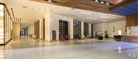 A spacious lobby with a polished marble floor, wooden ceiling panels, and a seating area with red chairs.