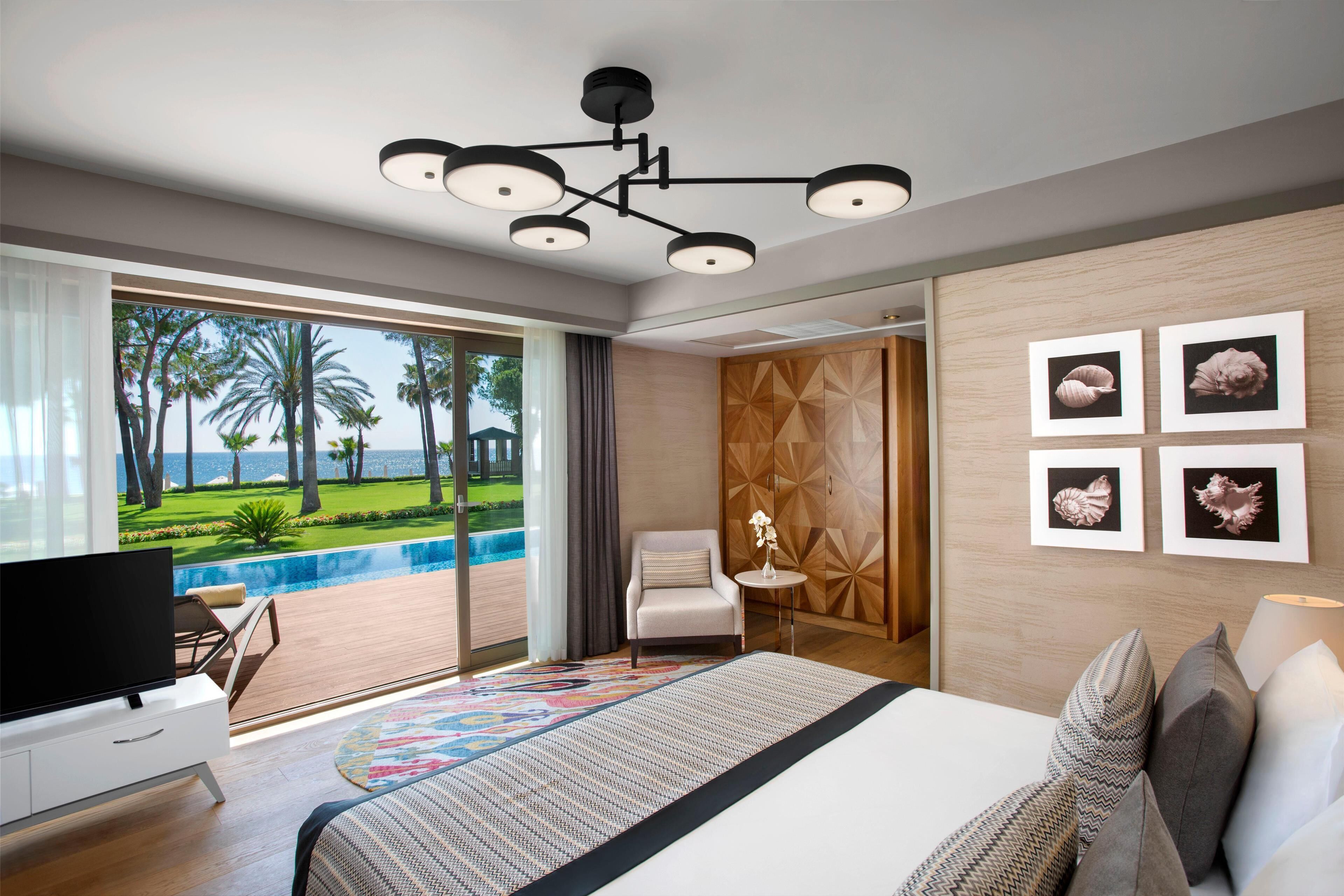 A modern hotel room with a large bed, a television, a view of a pool and palm trees, and artwork on the wall.