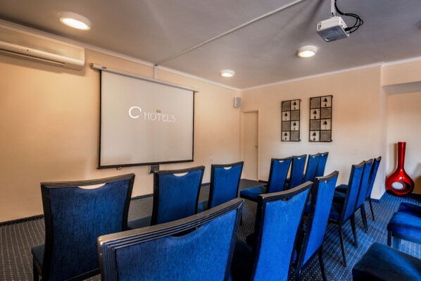 A conference room with a projector screen displaying "C HOTELS" logo, blue upholstered chairs, a red vase, and framed artwork on the wall. - Hacienda ForestView (Ma'alot - Tarshiha)