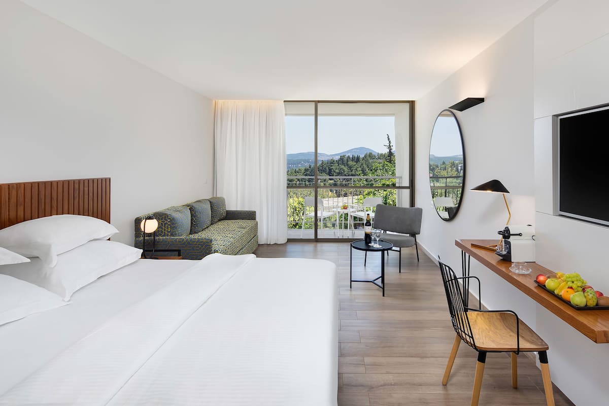A hotel room with a large bed, a sofa, a desk with a chair, a TV, and a view of the outdoors.
