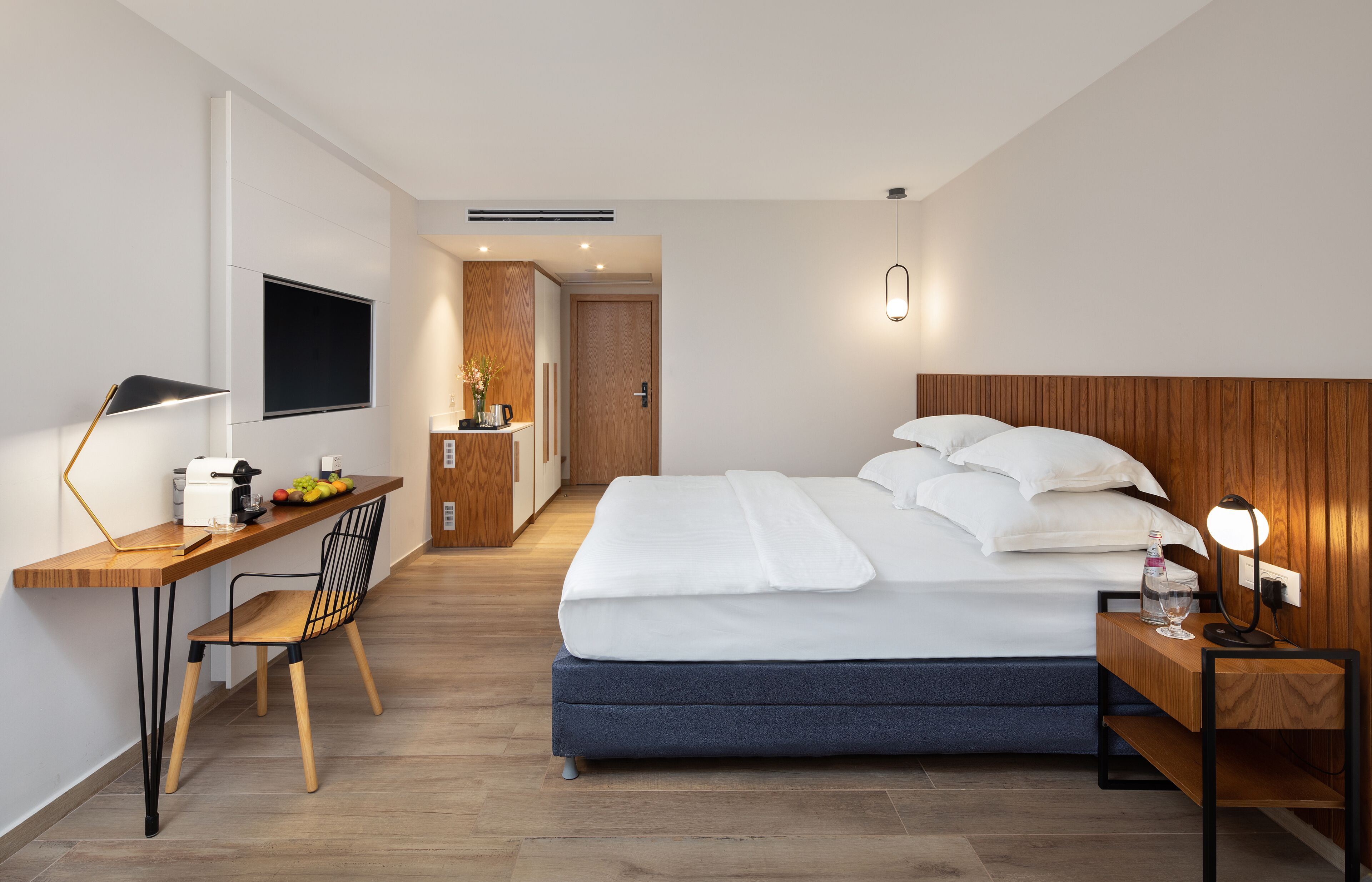 A modern hotel room with a bed, bedside table, lamp, and a desk with a chair.