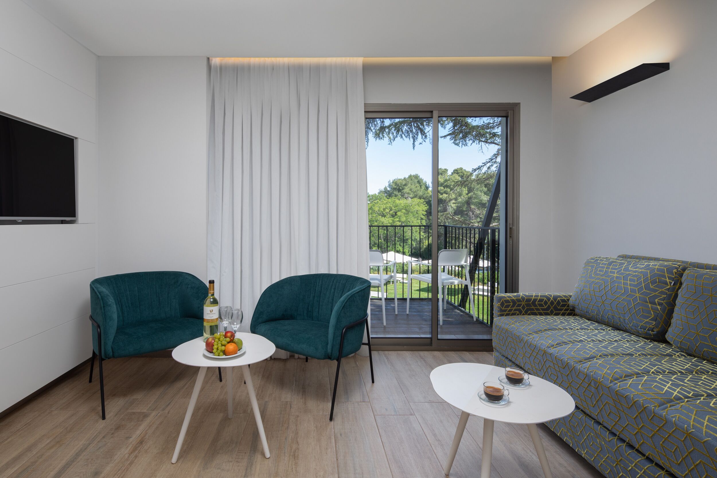 A modern living room with two teal armchairs, a patterned sofa, and a balcony with outdoor furniture.