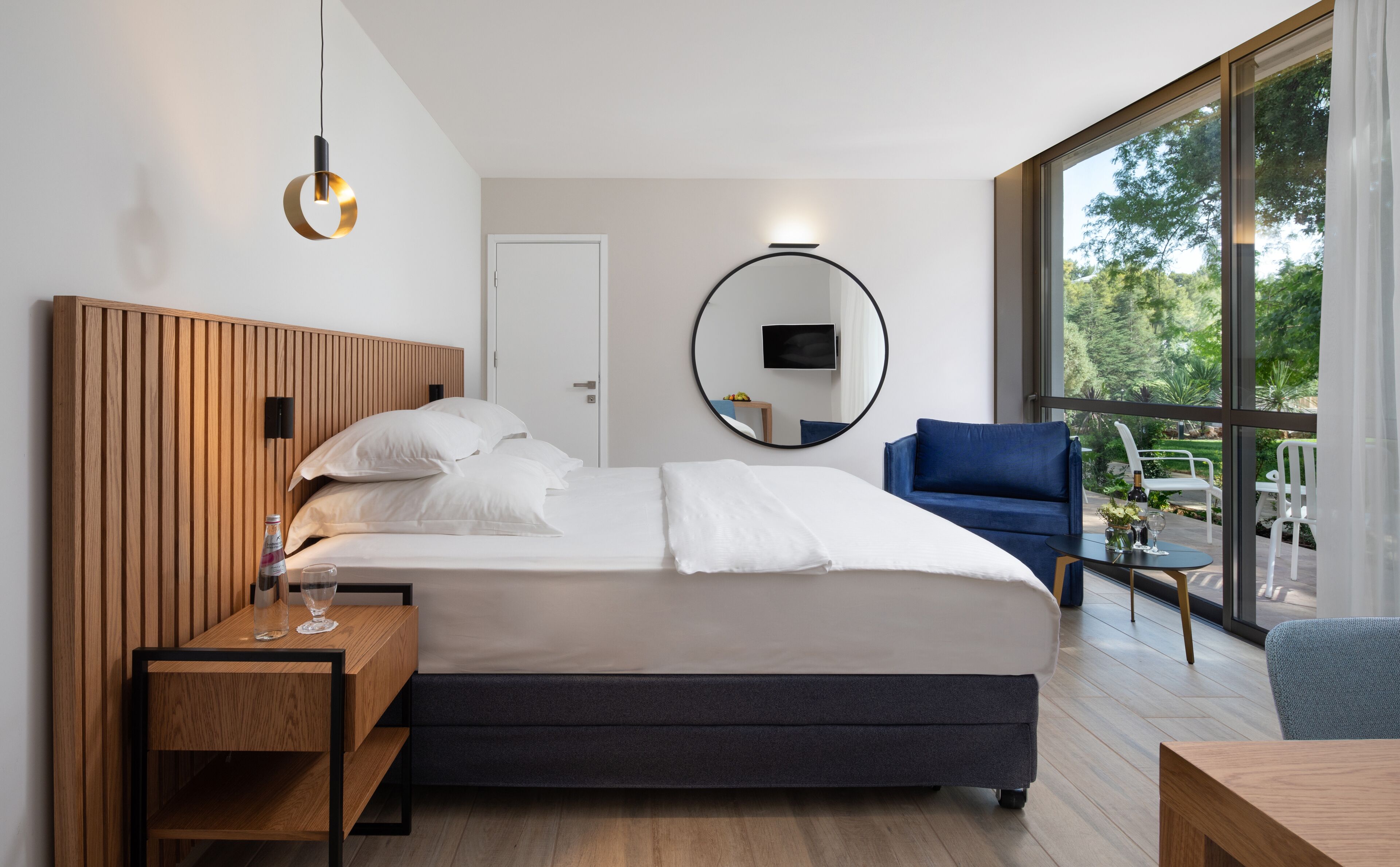 Habitación de hotel moderna con cabecera de madera, cama grande con sábanas blancas, espejo redondo, sillón azul y vistas a zonas verdes a través de amplios ventanales.