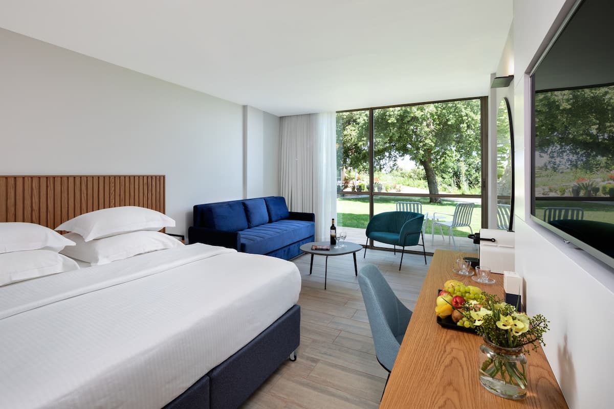 A hotel room with a bed, a blue sofa, a small table with a vase of flowers, and a view of a garden through large windows.