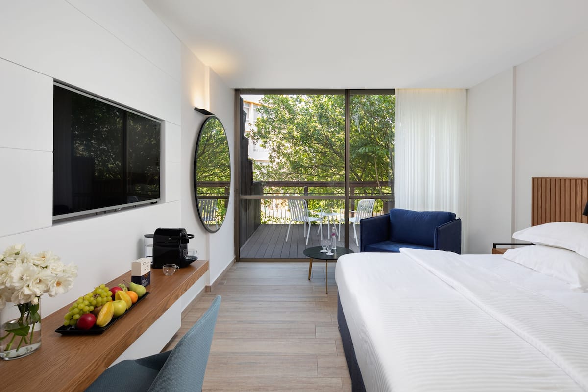 A modern hotel room with a large bed, a seating area with a blue sofa, a dining table with a coffee machine, and a large window with a view of trees.
