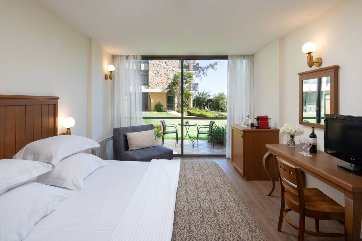 A hotel room with a bed, a desk, a television, and a view of the garden through the glass door.