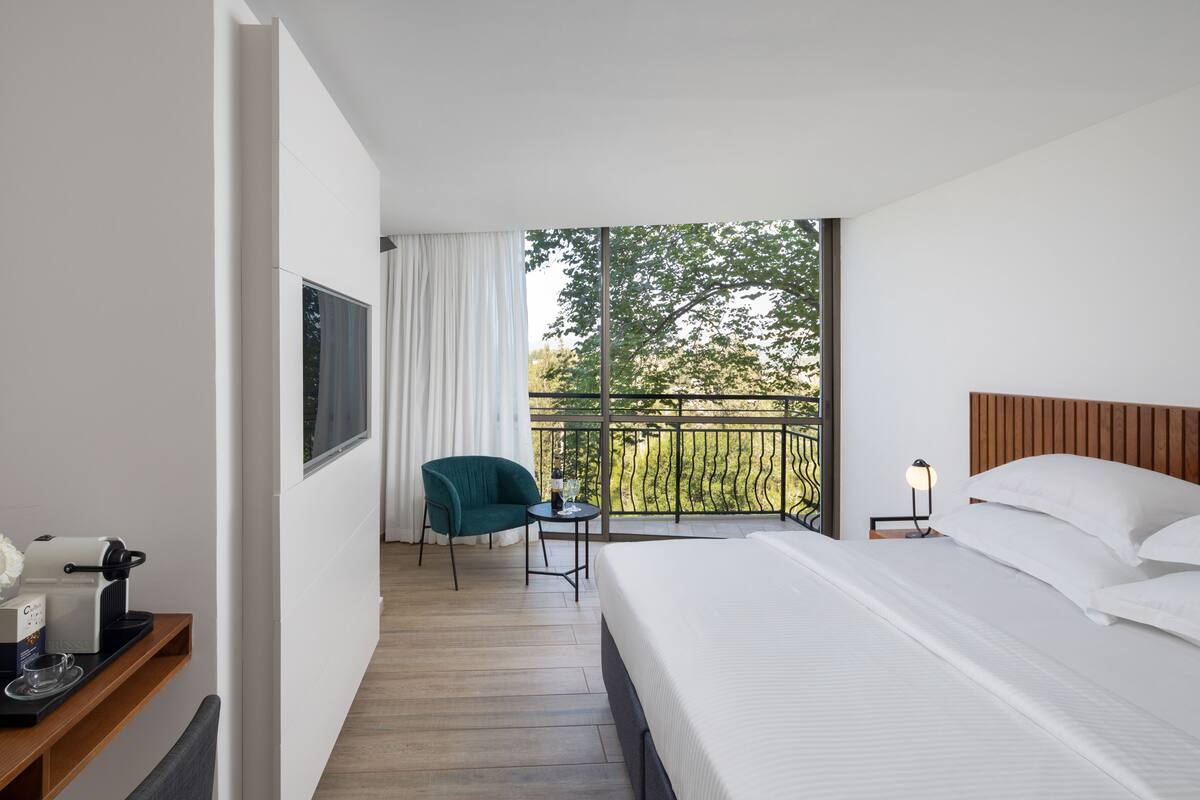 A hotel room with a bed, a small table with a bottle and glass, a chair, a coffee machine, and a view of a balcony with trees.