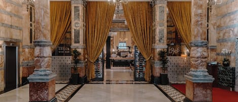 Un amplio vestíbulo con columnas de mármol, piso de diseño ajedrezado y una alfombra roja que conduce a una zona de bar.