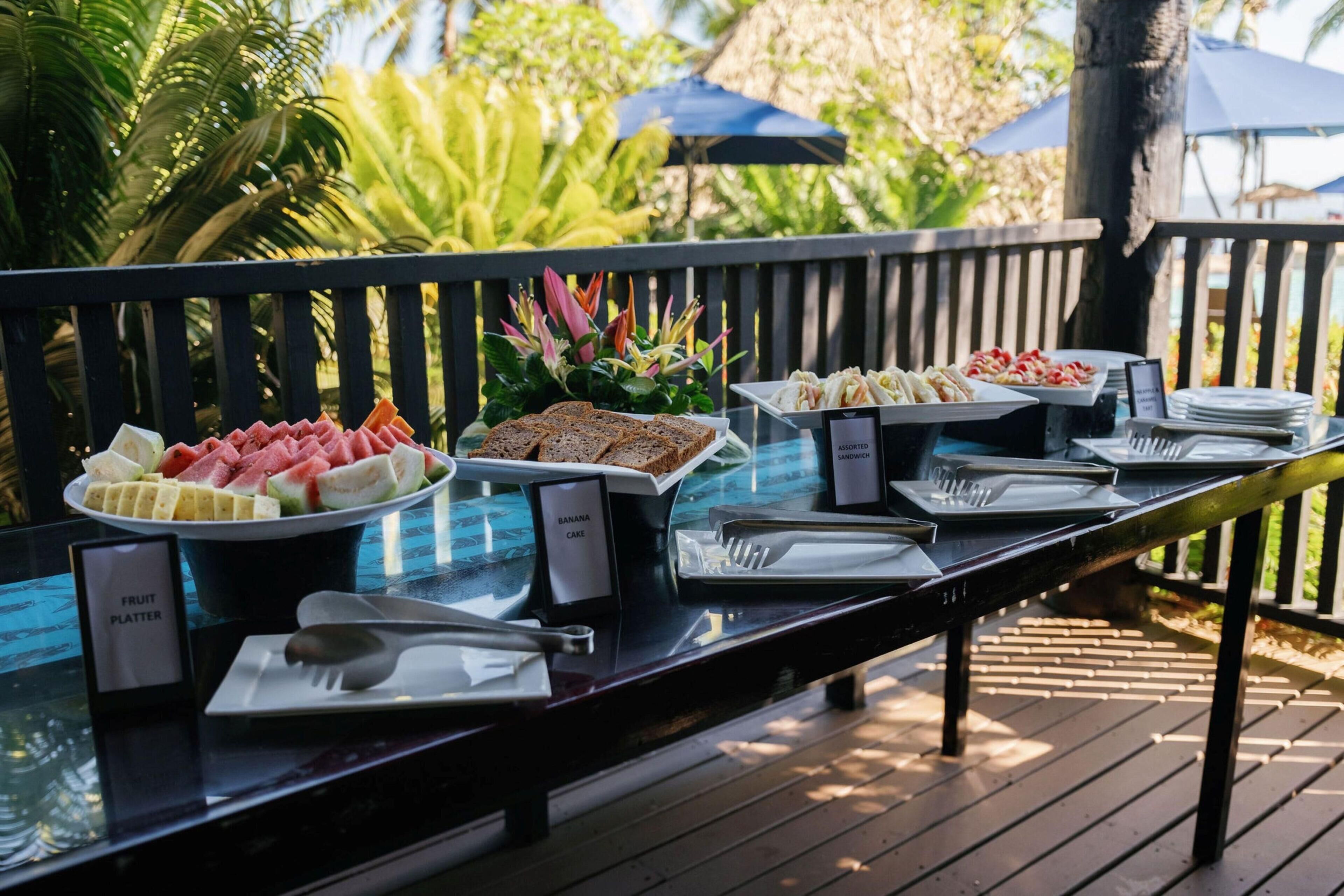 Een buffettafel met diverse gerechtjes, waaronder een fruitplateau, bananentaart en verschillende soorten belegde broodjes.