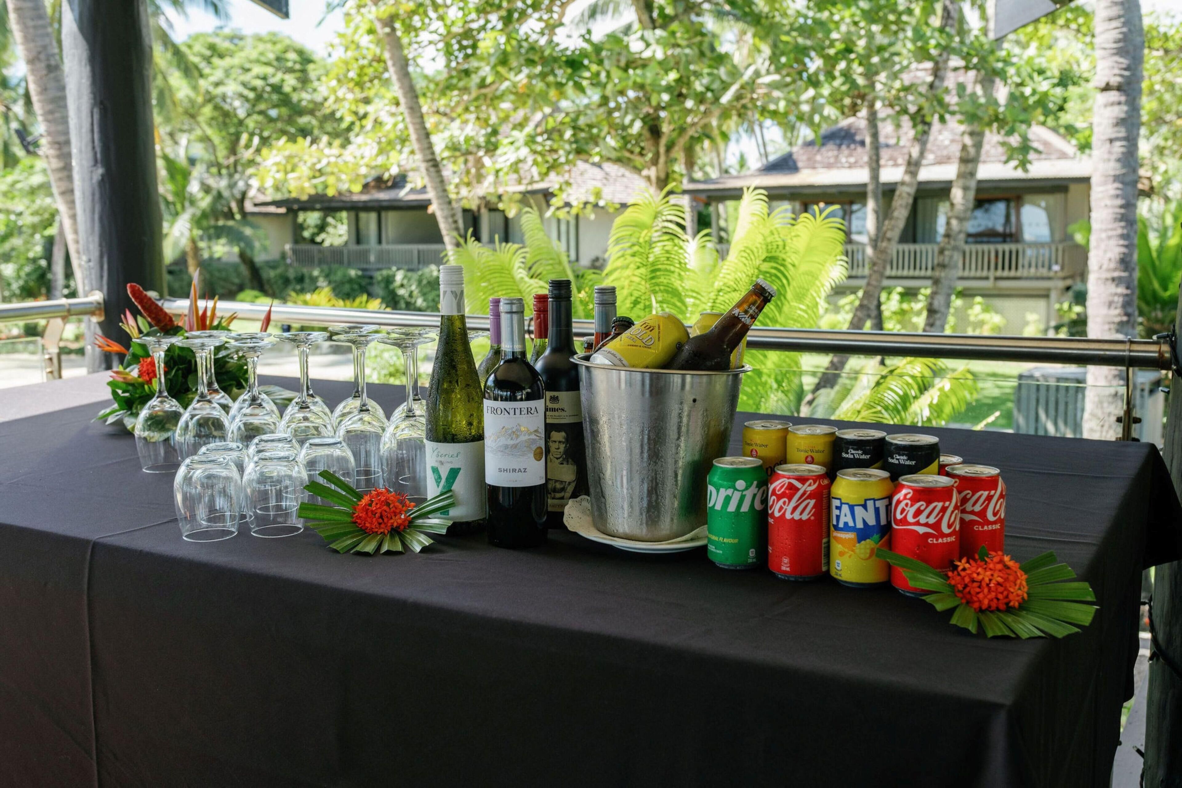 Une table dressée avec des bouteilles de vin, des verres et des canettes de soda, offrant une vue sur un paysage tropical.