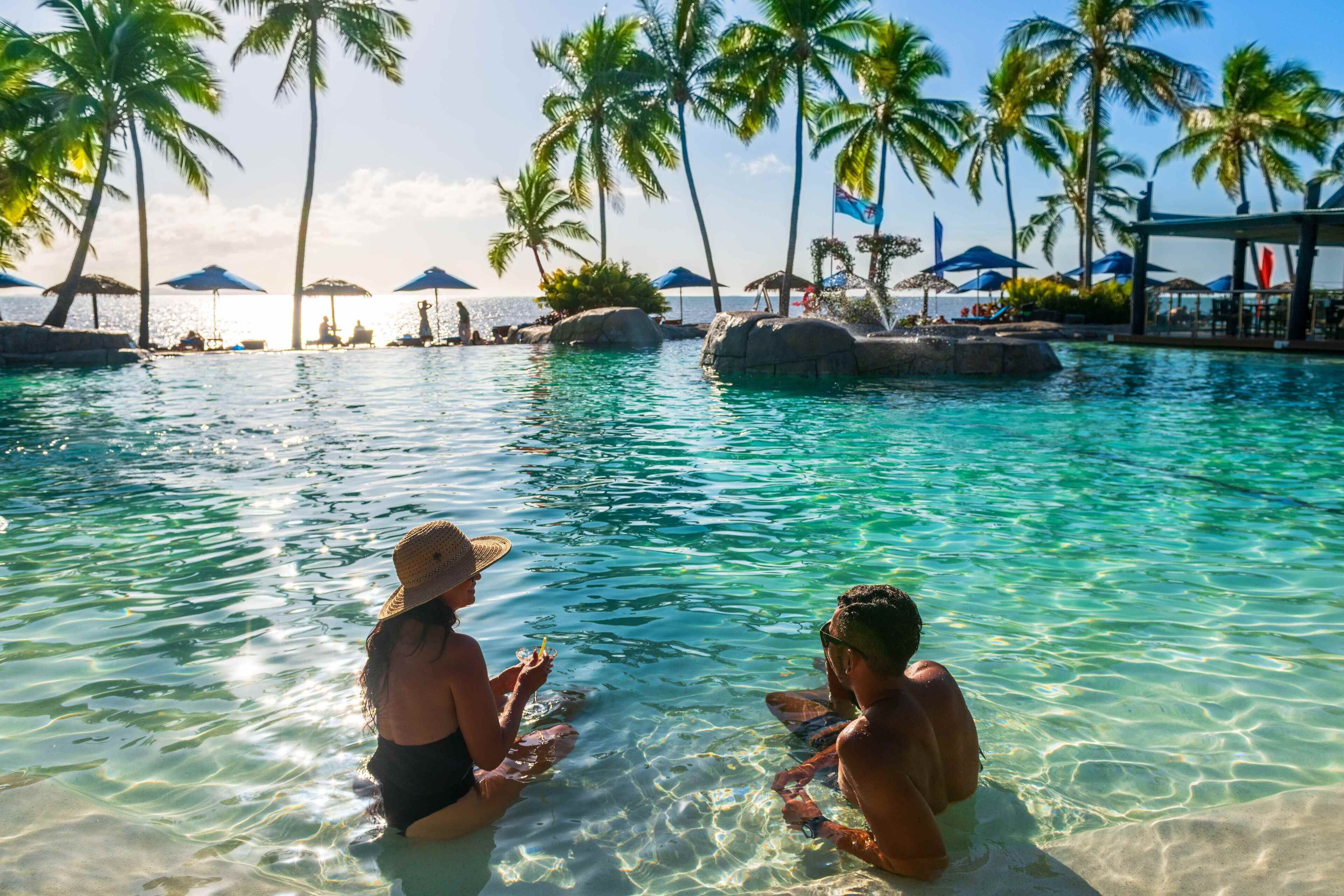 A clear pool with palm trees and umbrellas, surrounded by rocks and a lounge area.