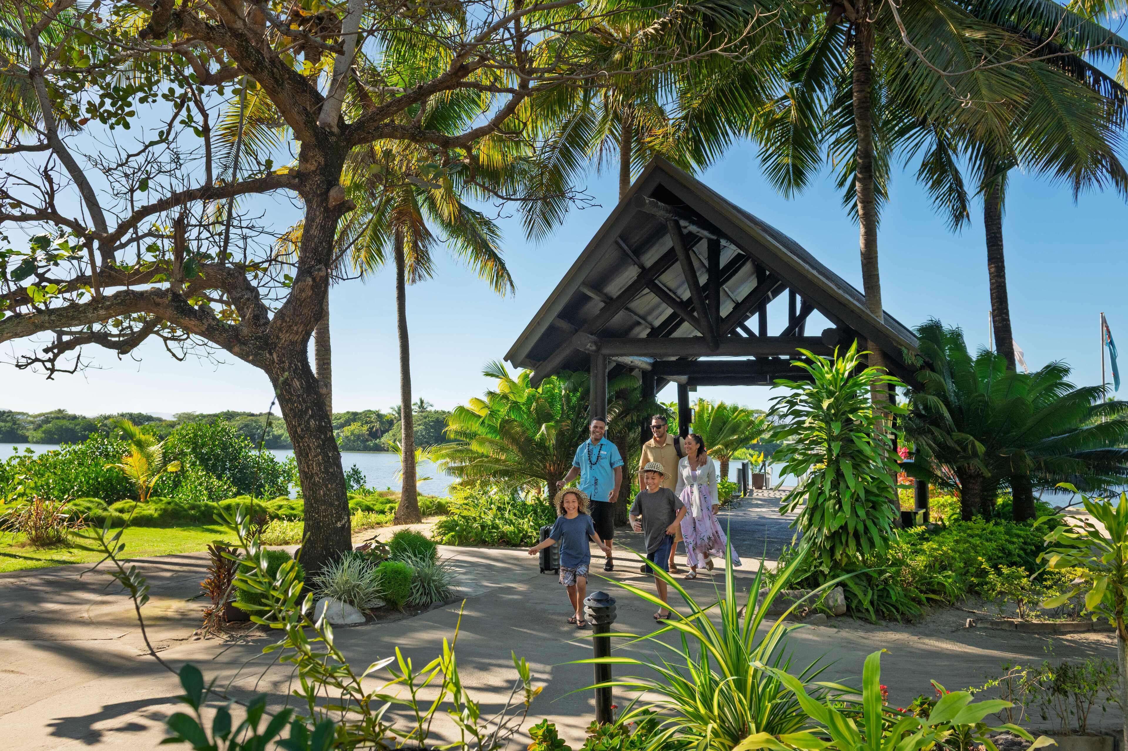 A covered walkway with a metal roof, surrounded by lush greenery and palm trees, leading to a body of water.