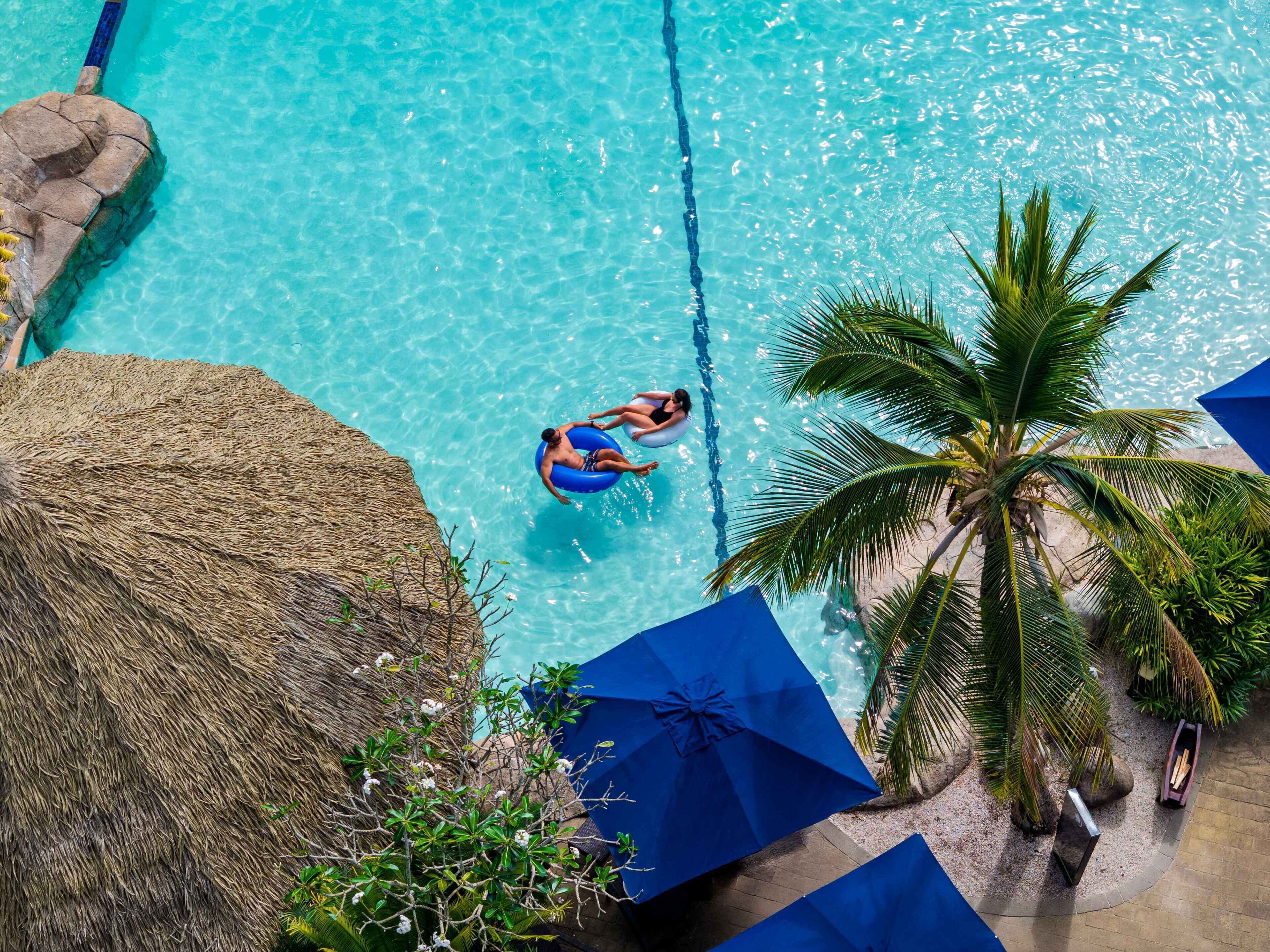 A clear blue pool with a person floating on an inflatable ring, surrounded by palm trees and blue umbrellas.
