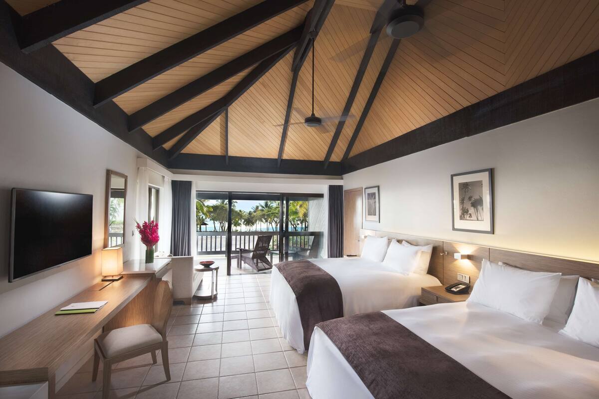 A hotel room with two beds, a desk, a TV, and a balcony with a view of palm trees.