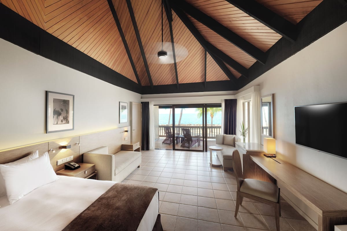 A hotel room with a bed, a desk, a TV, and a balcony with a view of the sea.