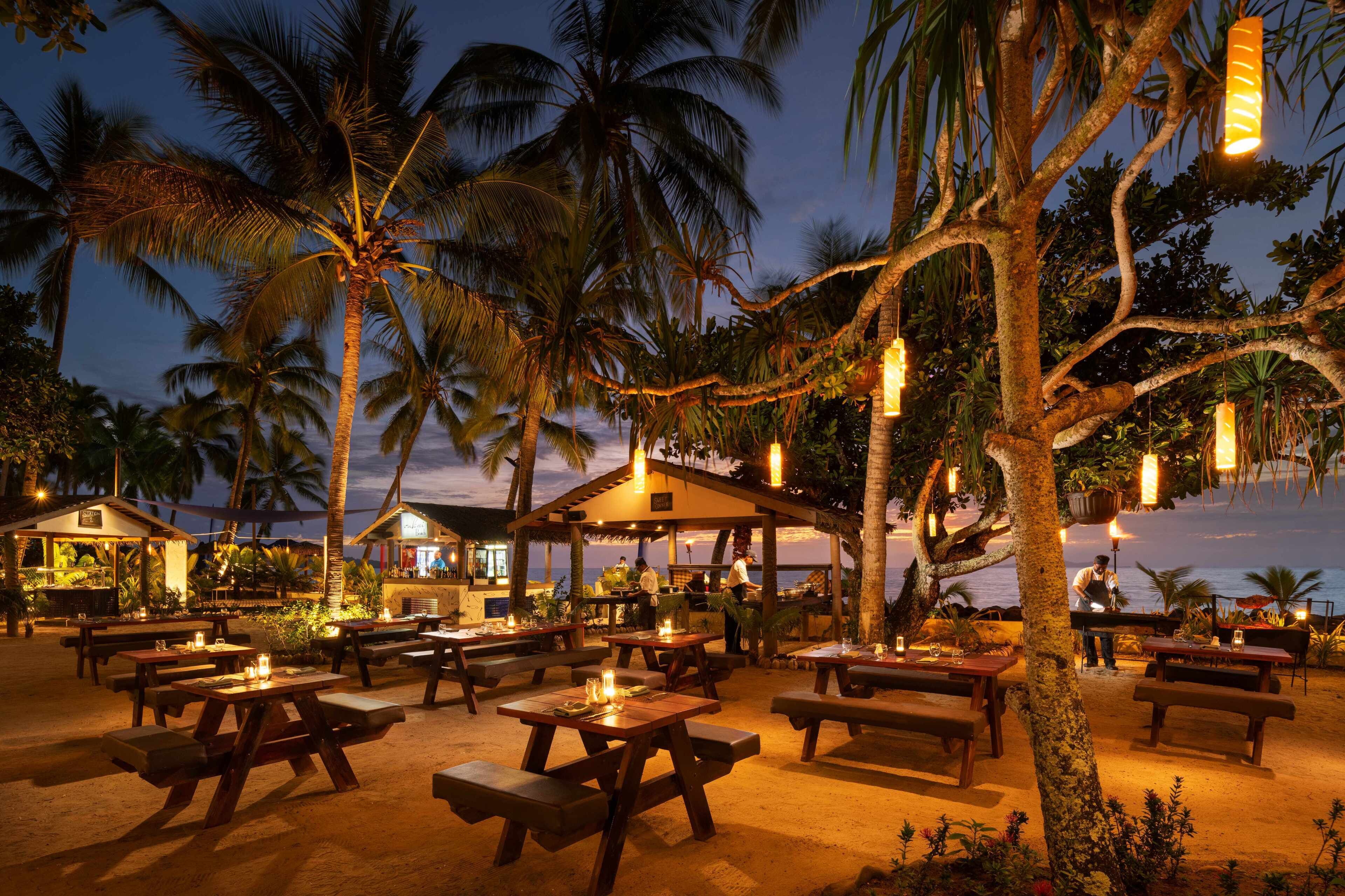 An outdoor dining area with wooden tables and benches, surrounded by palm trees and string lights, overlooking a beach at dusk.