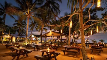 An outdoor dining area with wooden tables and benches, surrounded by palm trees and string lights, overlooking a beach at dusk.