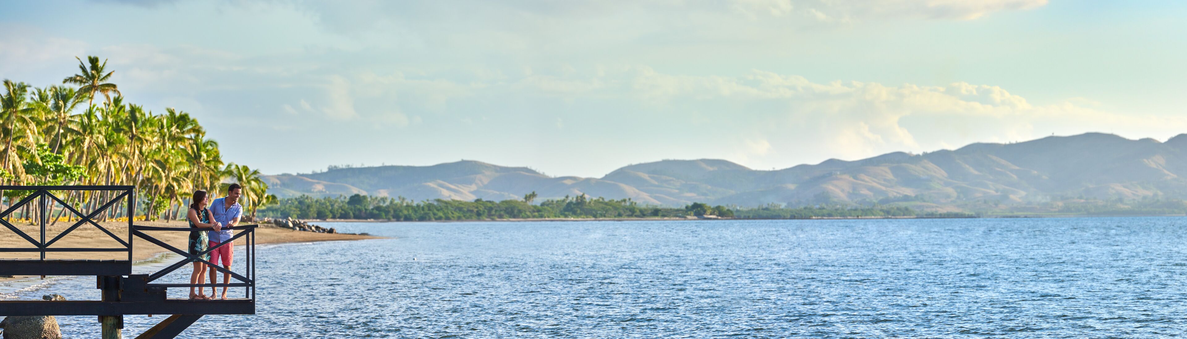 A wooden pier with a metal railing, overlooking a calm body of water with palm trees and mountains in the background.