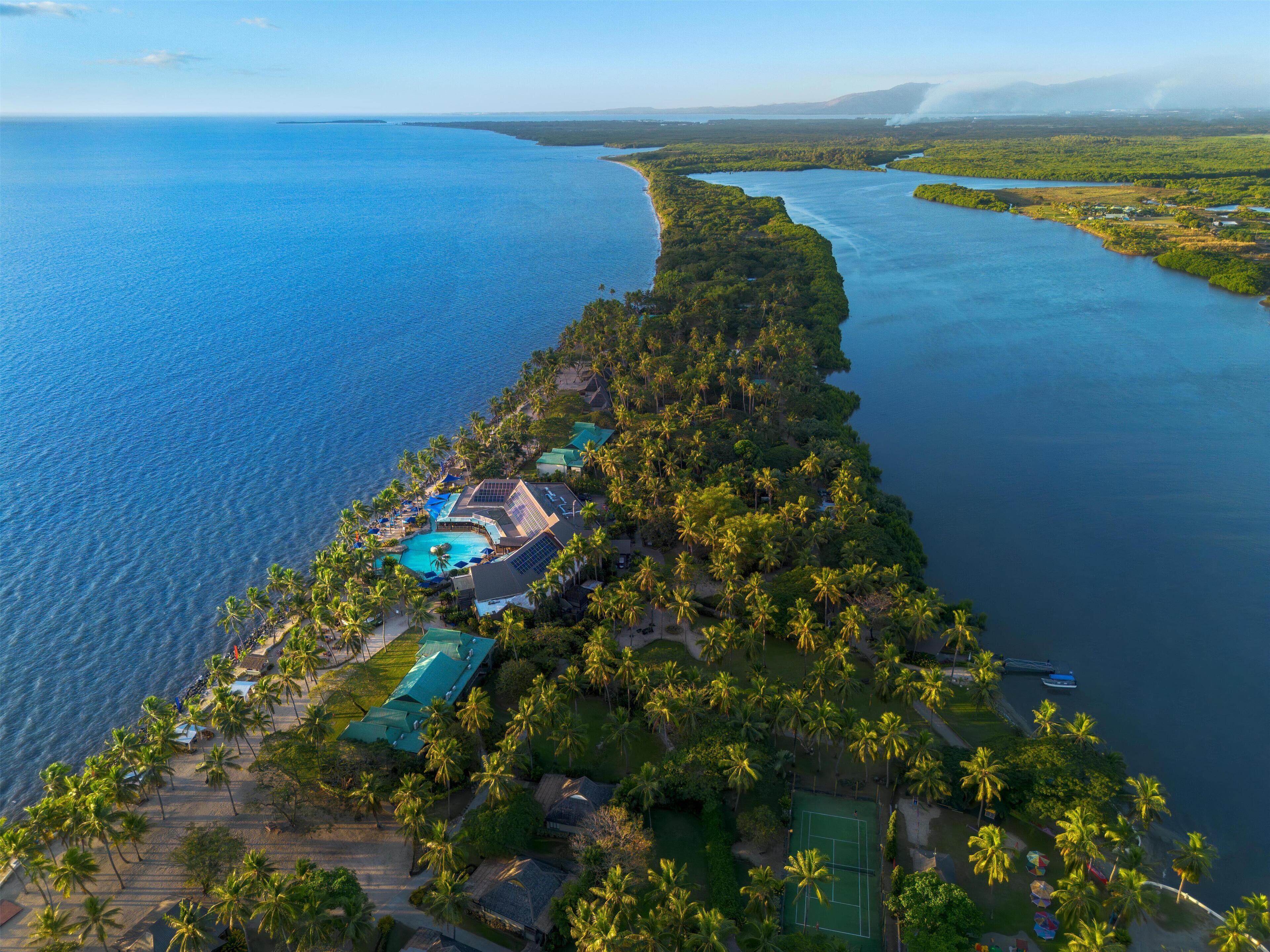 An aerial view of a coastal resort with a swimming pool, tennis court, and lush greenery.