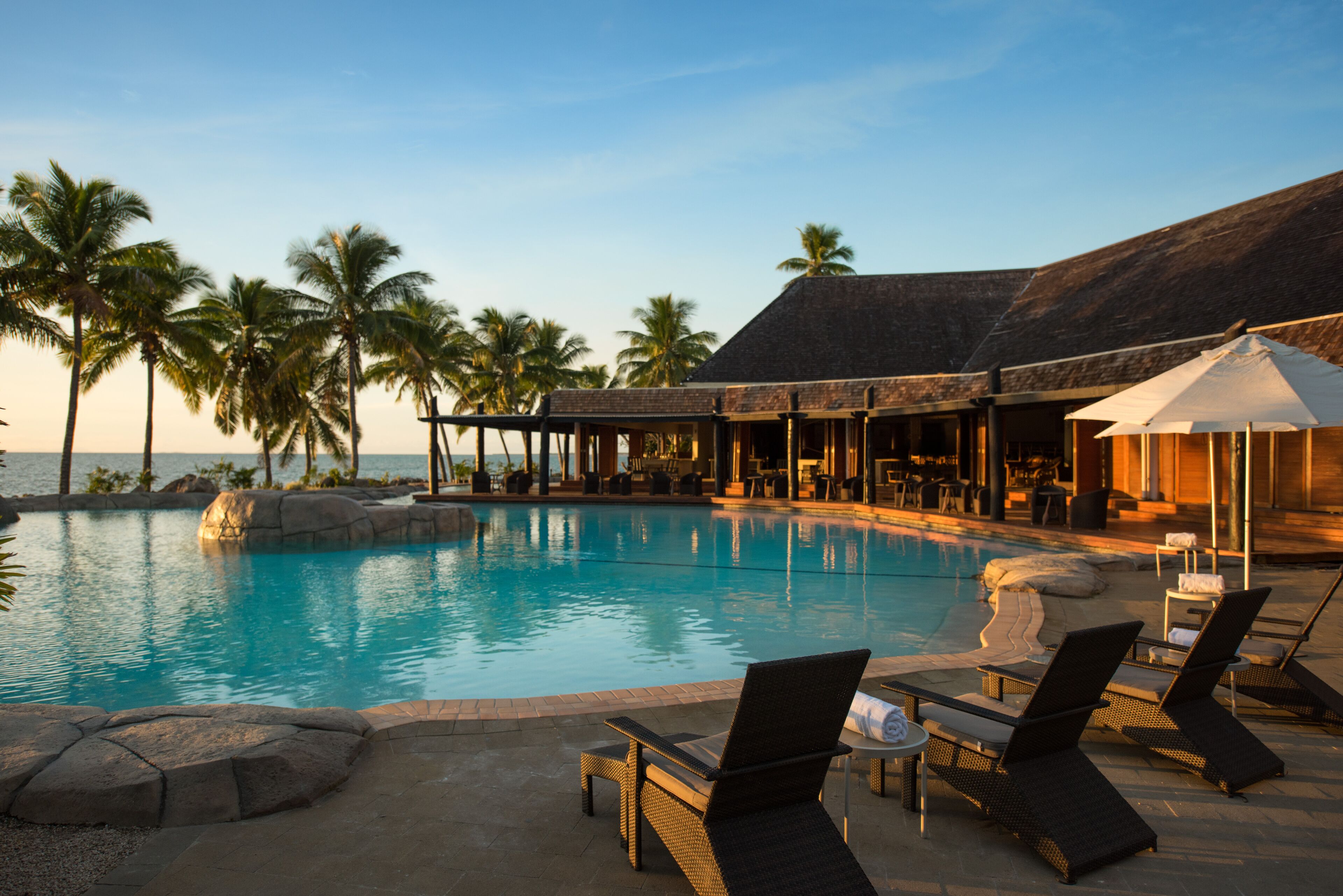 A pool area with lounge chairs and umbrellas, surrounded by palm trees and a building with a thatched roof.