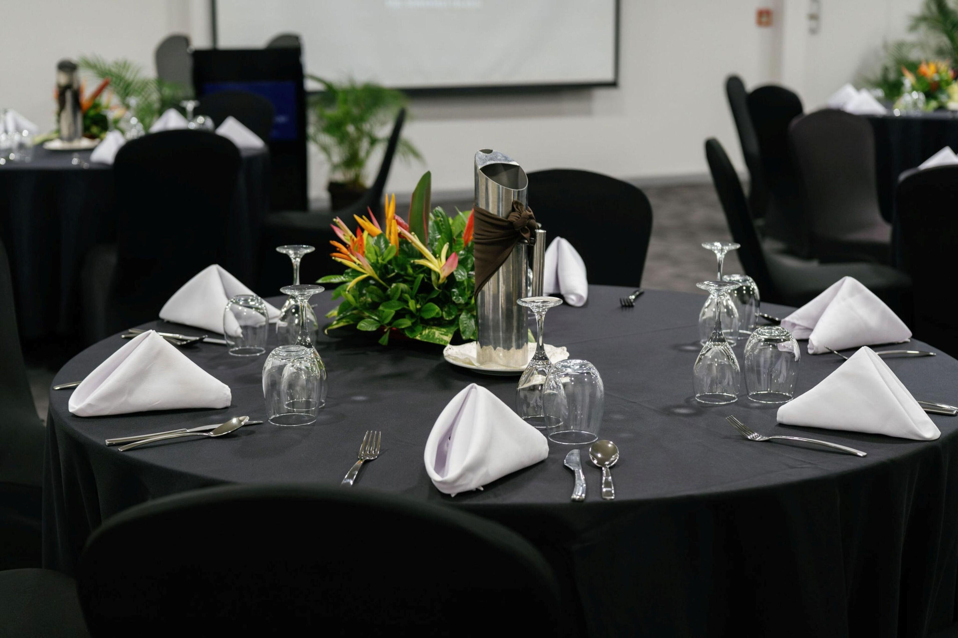 Une salle de conférence avec des tables rondes disposées pour une réunion, équipée de marque-places, de verres et d’un centre de table décoratif.