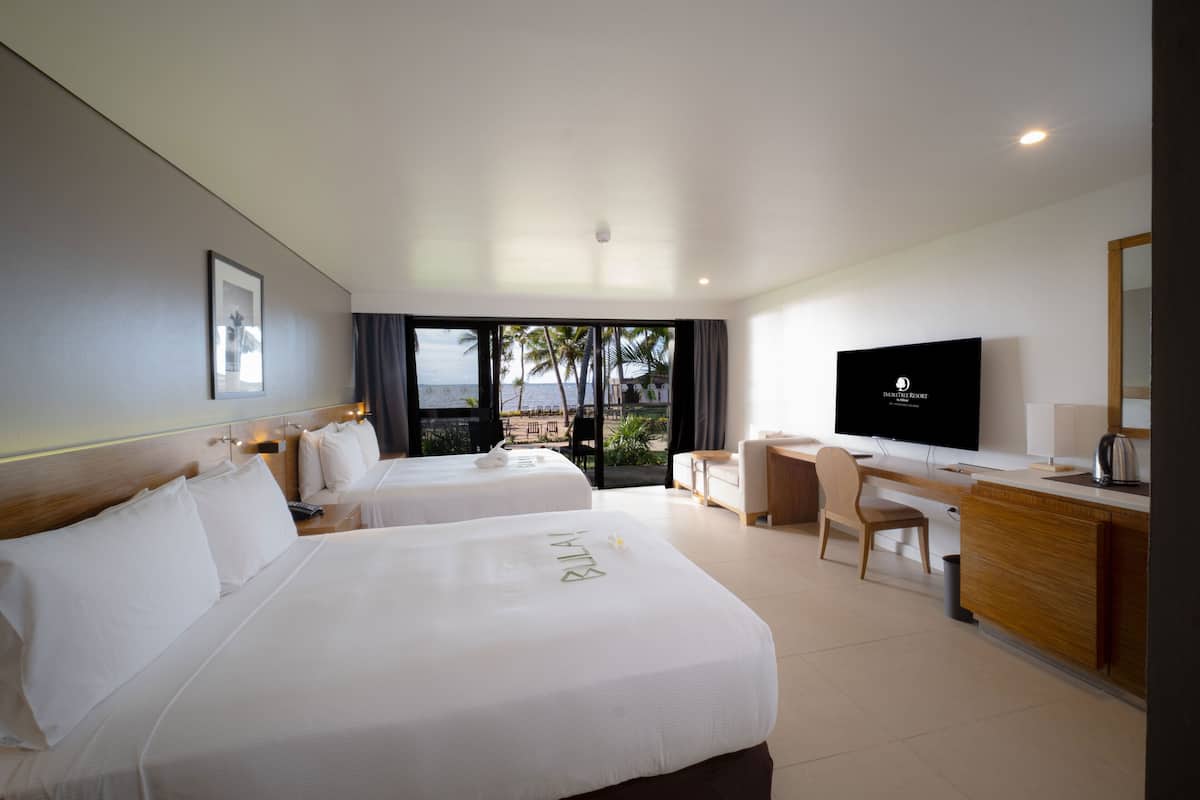 A hotel room with two beds, a desk, a TV, and a view of the ocean.