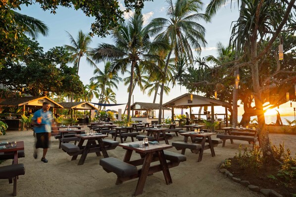 Un área de comedor al aire libre con mesas y bancos de madera, rodeada de palmeras y vistas a la playa.