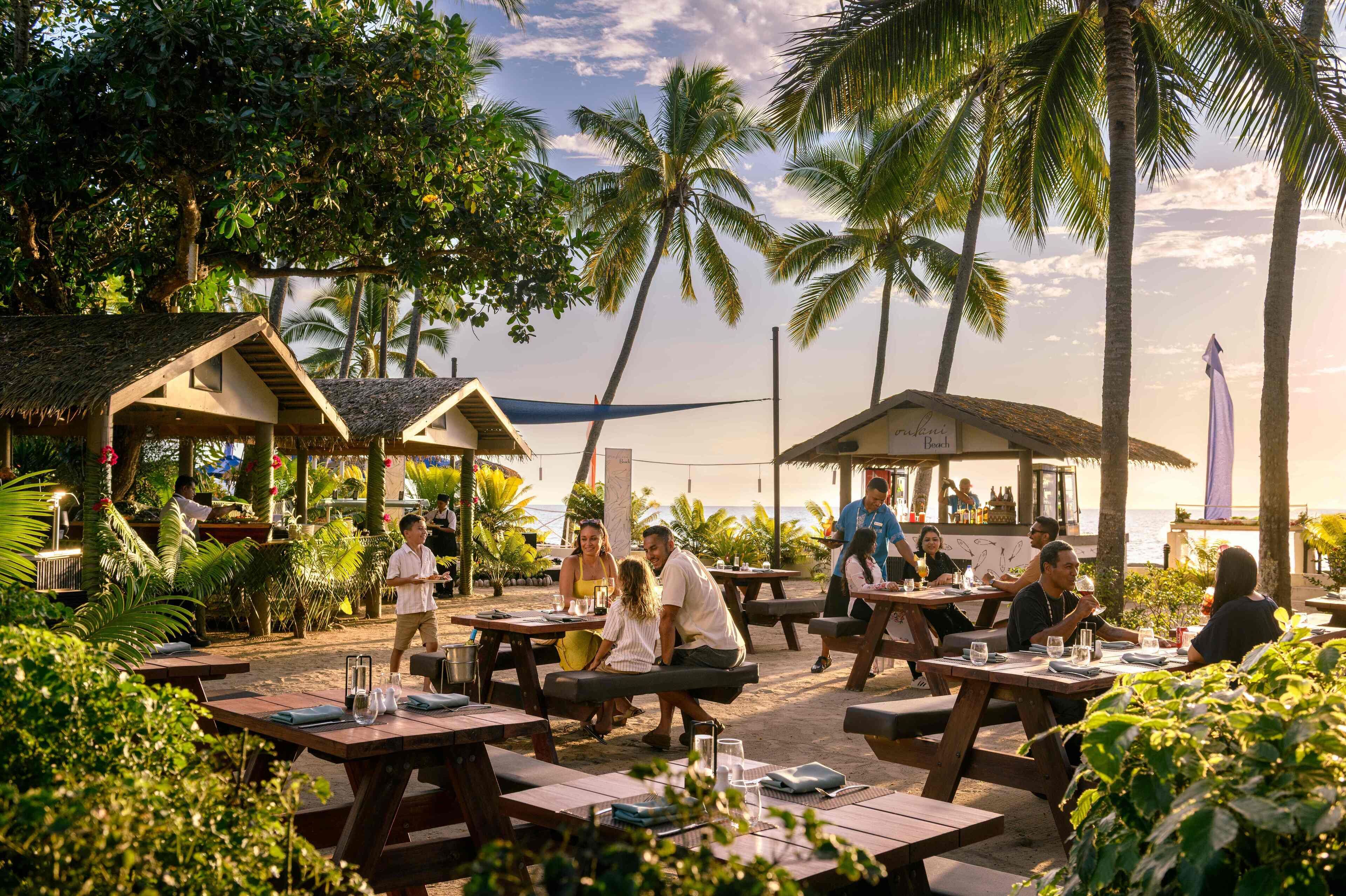 A beachside restaurant with outdoor seating, palm trees, and a view of the ocean.