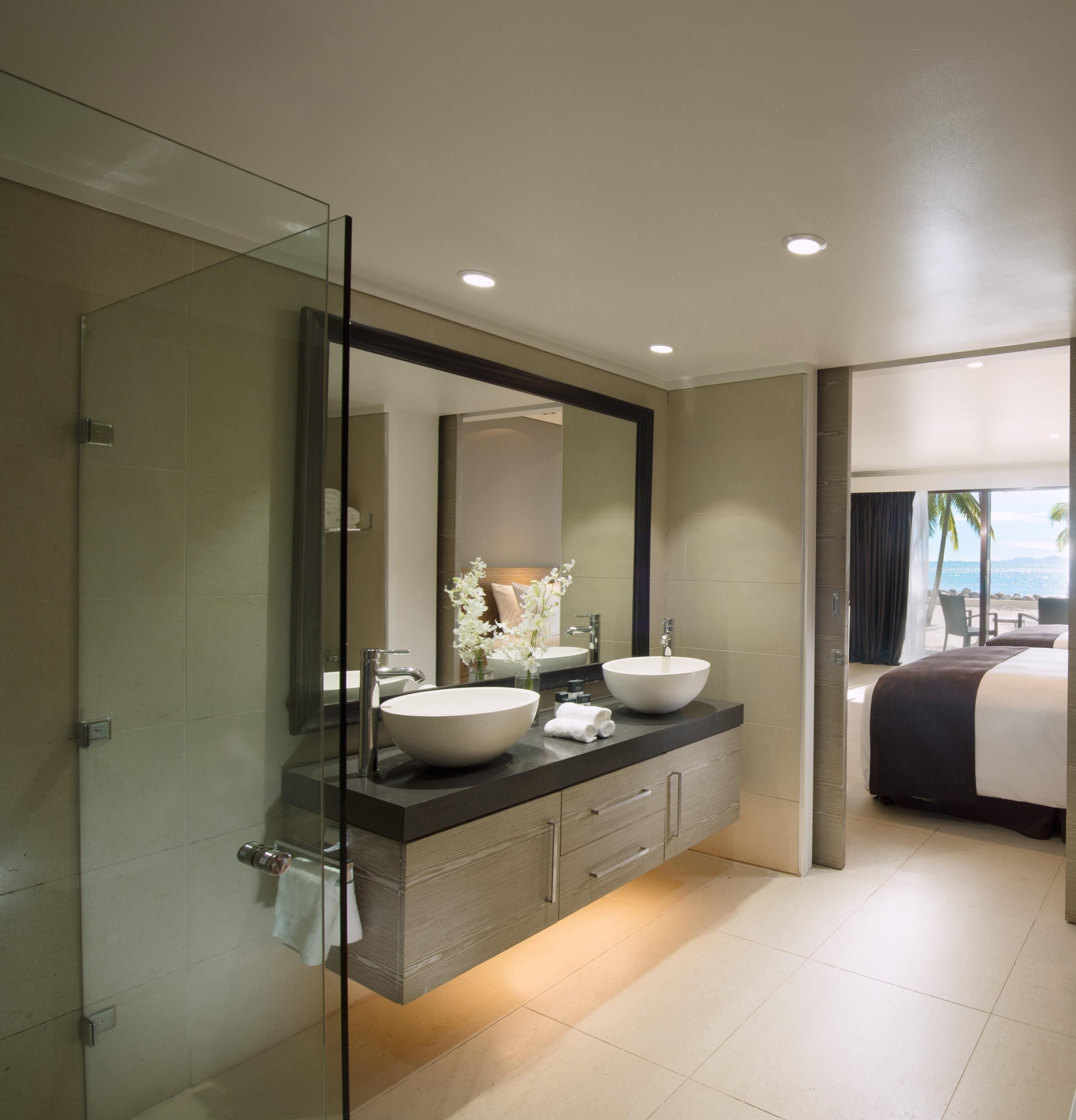 A modern bathroom with a glass shower enclosure, a large mirror, dual sinks, and a view of the beach.