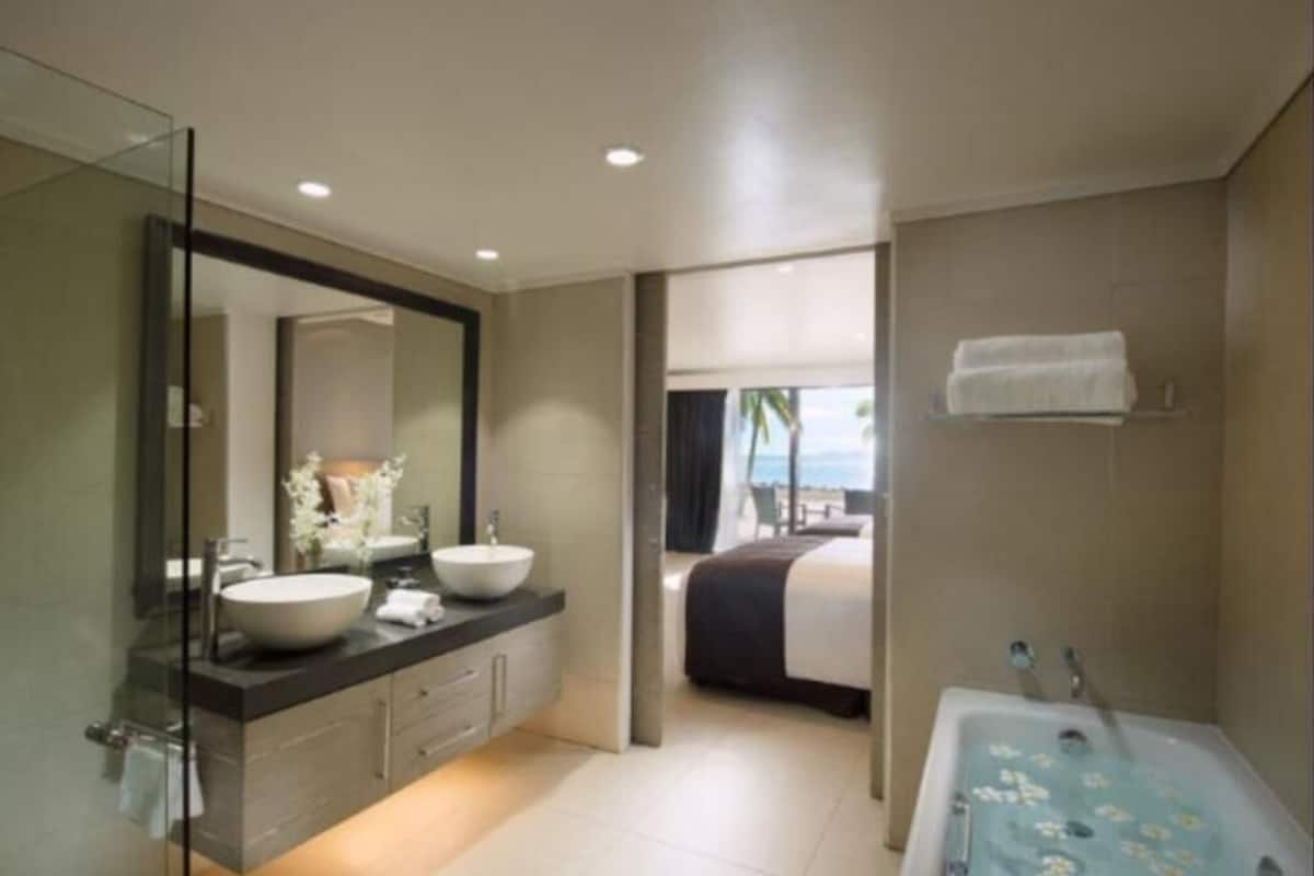 A modern bathroom with a large mirror, two sinks, a bathtub, and a view of a bedroom with a bed and a window.