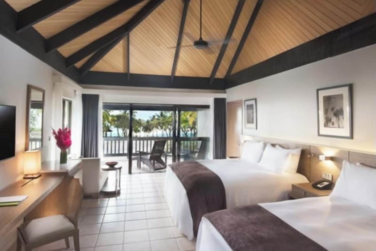 Une chambre d’hôtel avec deux lits, un bureau, une chaise, une télévision et un balcon donnant sur des palmiers.