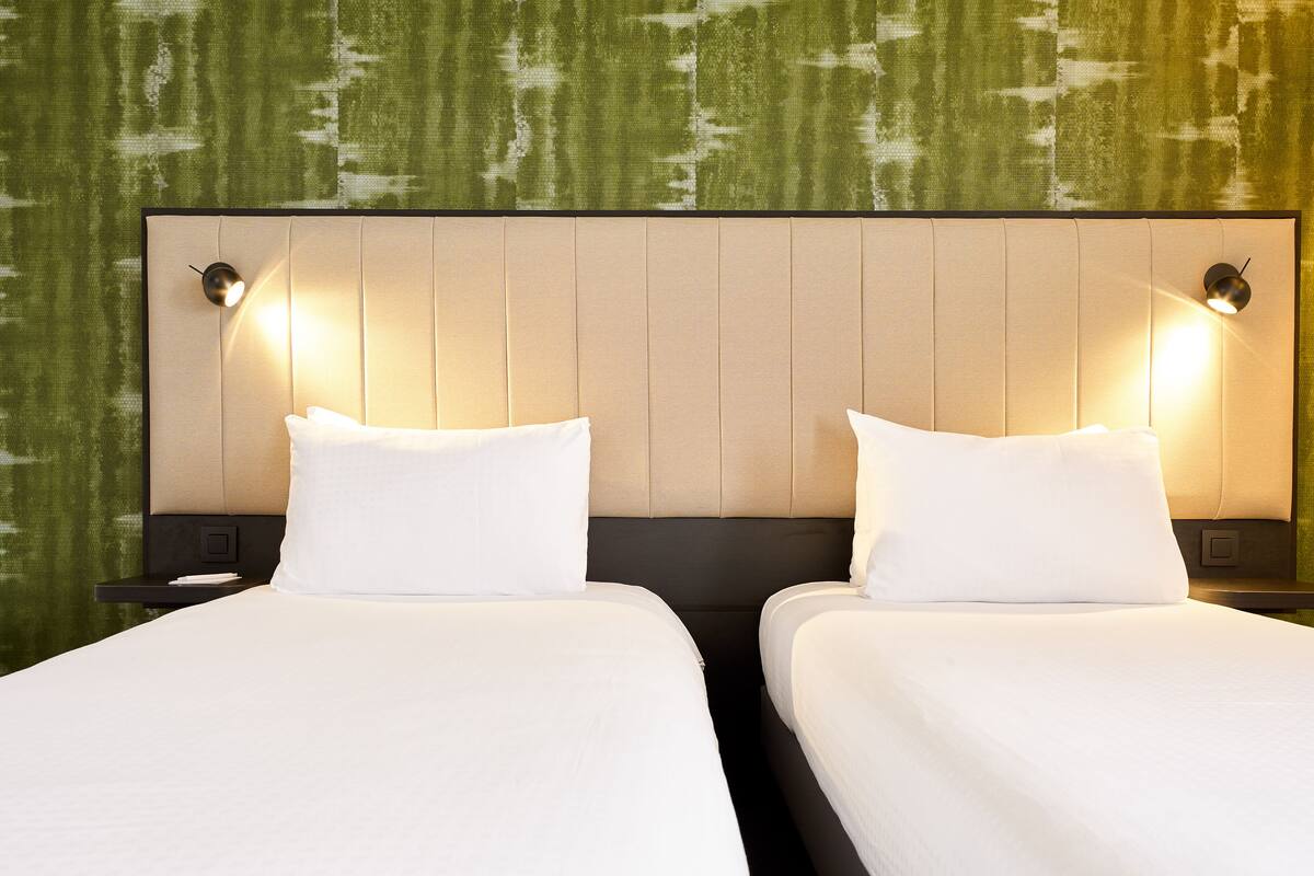 Two adjacent beds with white linens and headboards, each with a wall-mounted nightstand and a lamp.