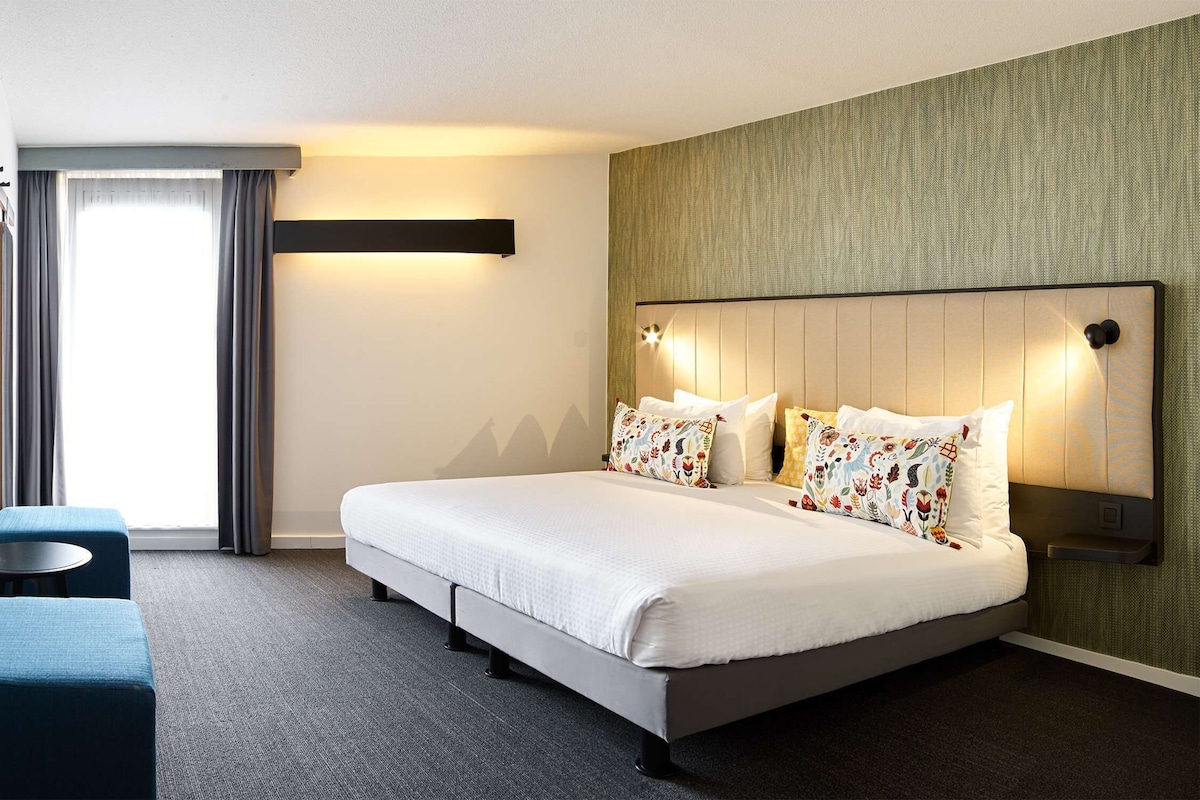 Una habitación de hotel con una cama, una mesita, un sofá y una ventana con cortinas.