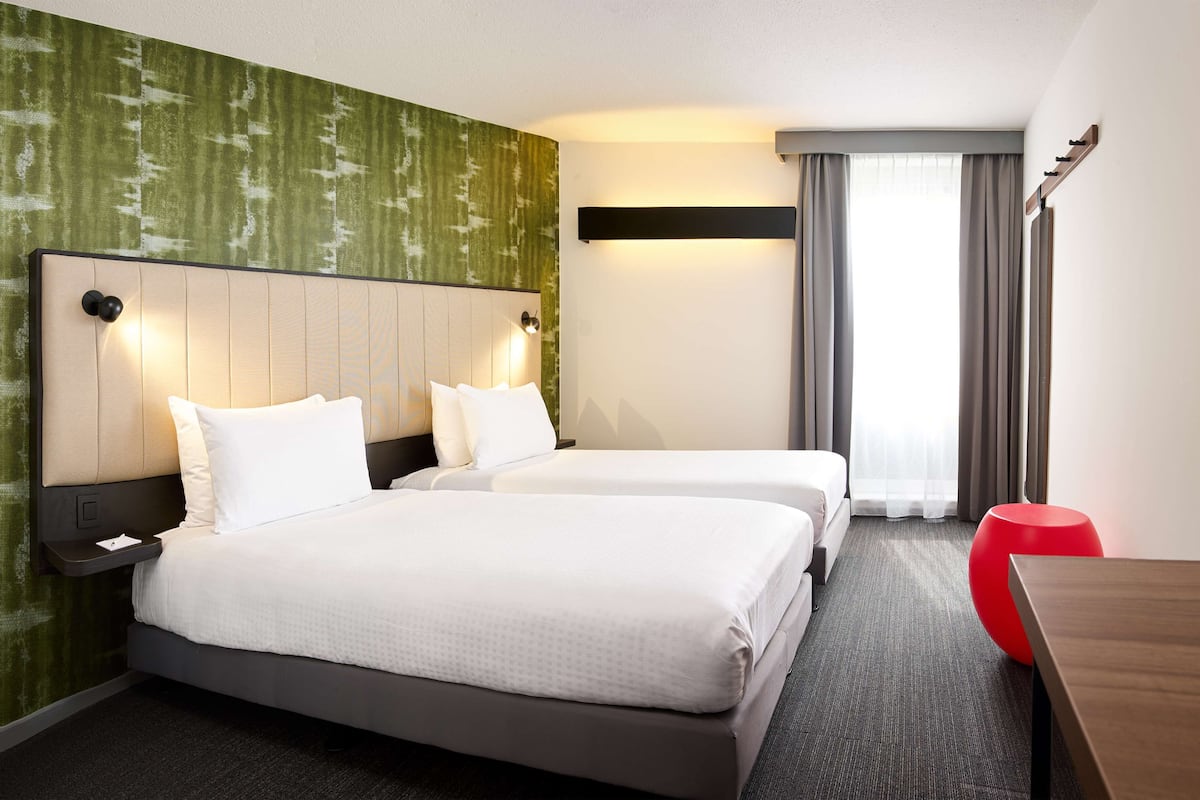 A hotel room with two beds, a red ottoman, and a large window with curtains.