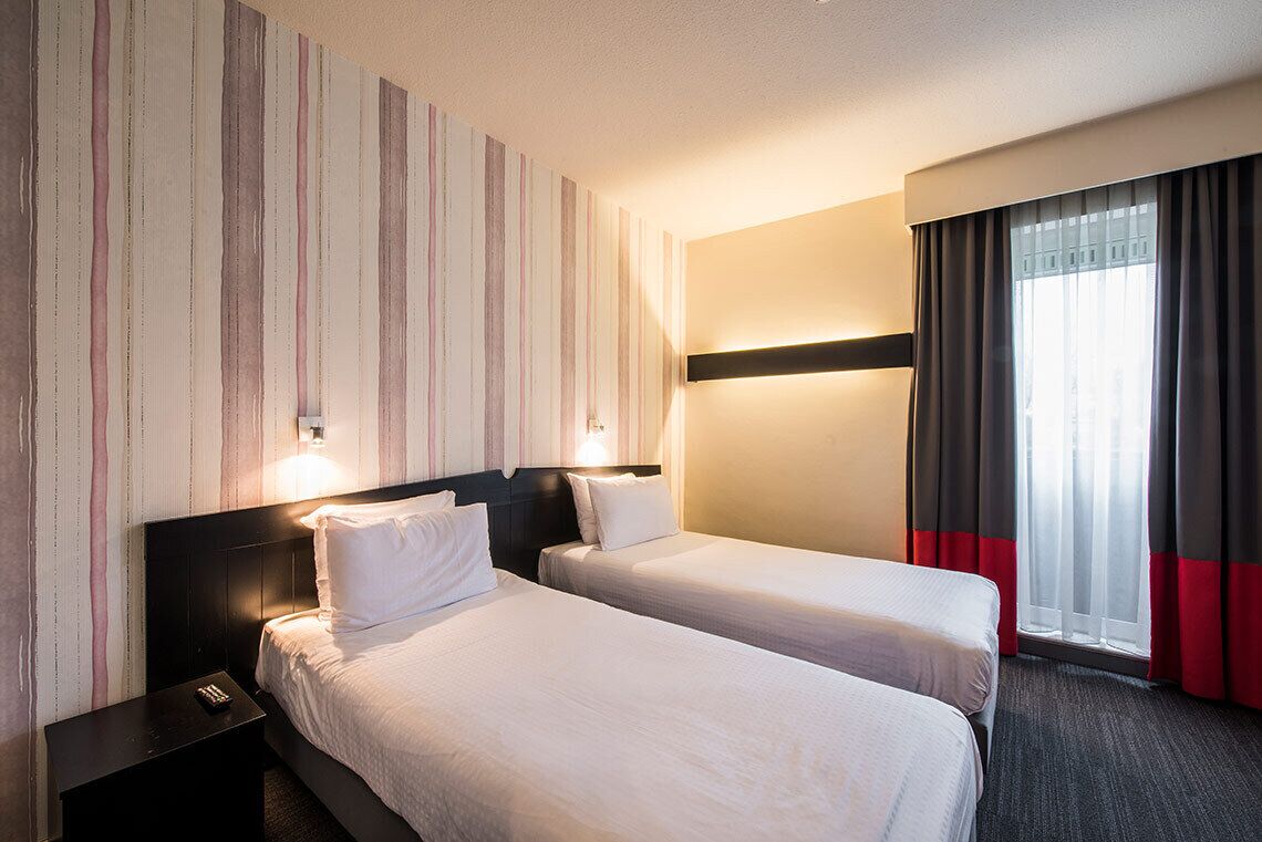 A hotel room with two beds, a nightstand, a wall-mounted light, and a window with curtains.