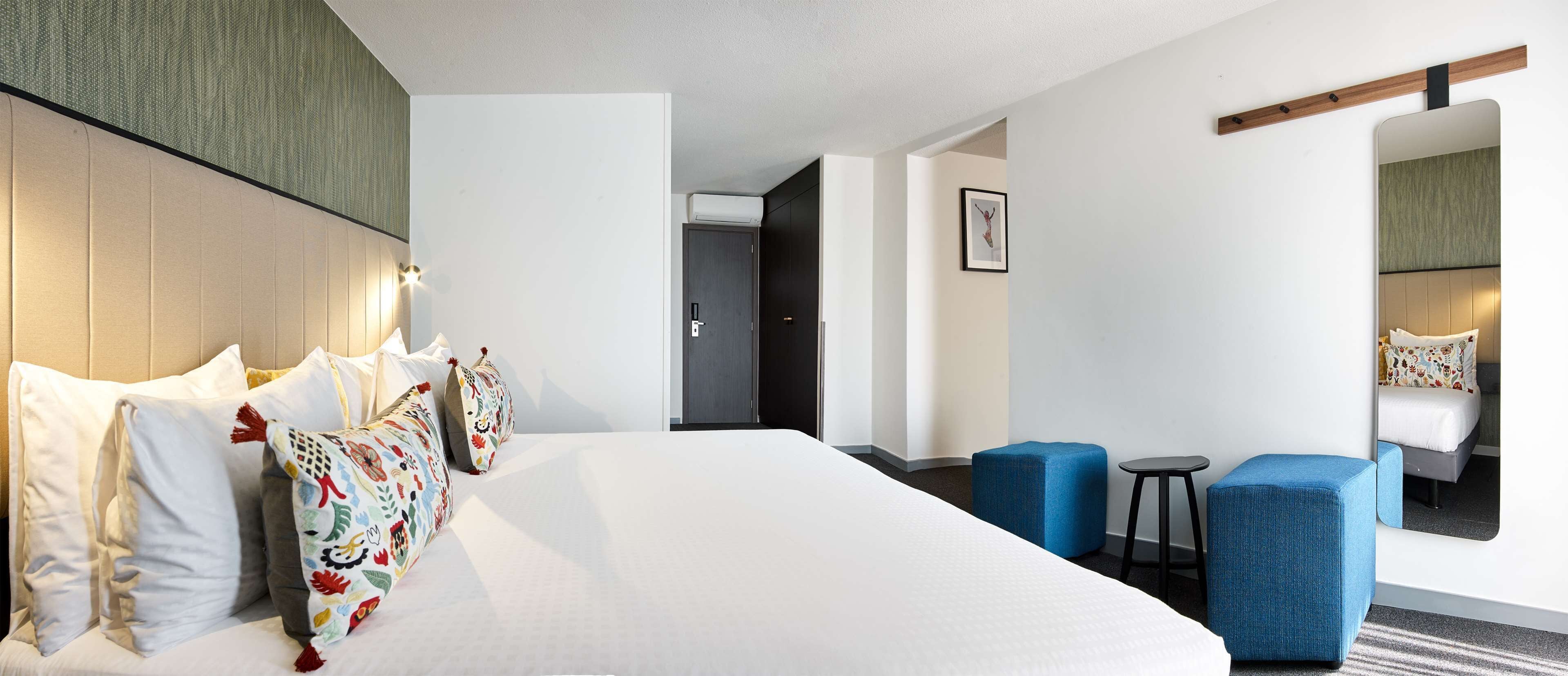 A hotel room with a large bed, two blue armchairs, a small table, and a mirror.