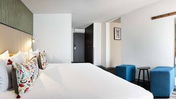 A hotel room with a large bed, two blue armchairs, a small table, and a mirror.