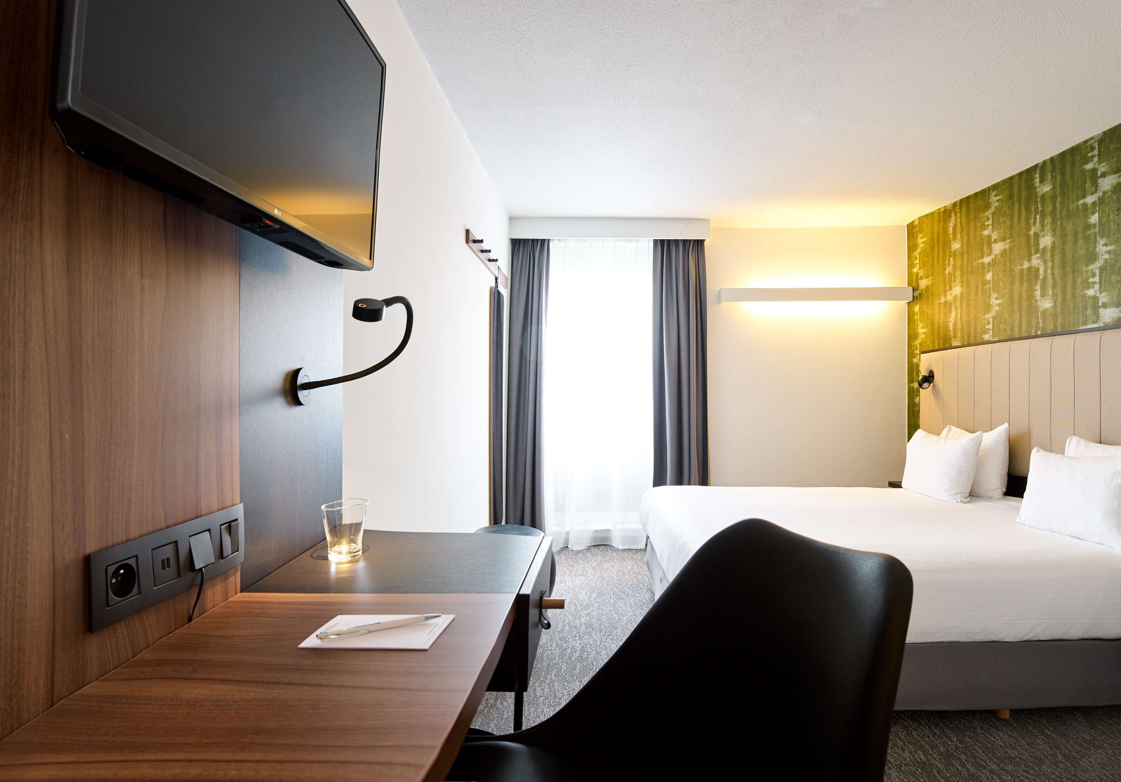 Habitación de hotel con cama, escritorio, silla, televisor y ventana con cortinas.