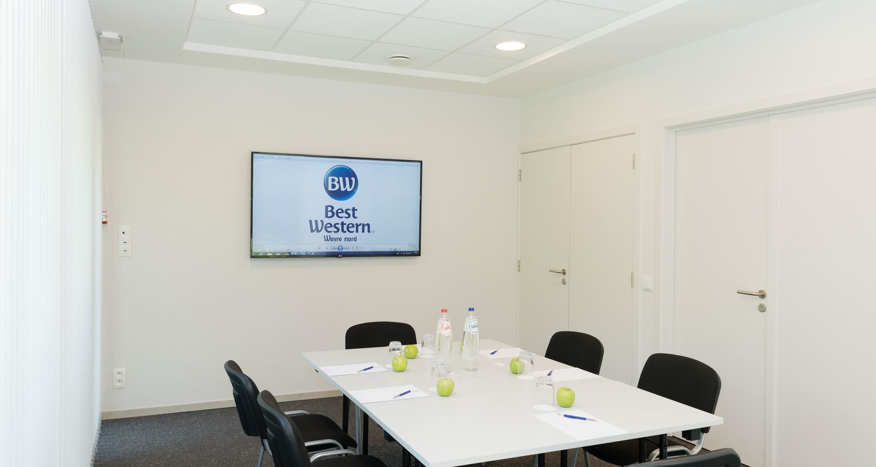 Une salle de conférence avec une table blanche, des chaises noires, un écran affichant le logo « BW Best Western », ainsi que quelques pommes et des bouteilles d’eau posées sur la table.