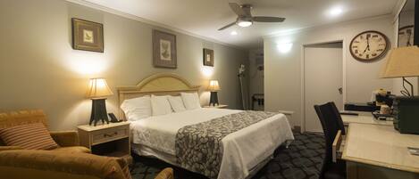 A hotel room with a bed, a desk, a chair, a lamp, a clock, and a ceiling fan.